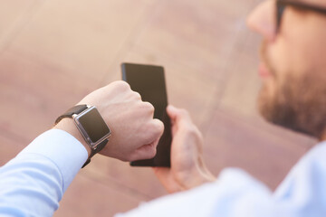 Young businessman holding smartphone and looking at his smartwatch while spending time outdoors, focus on male hand
