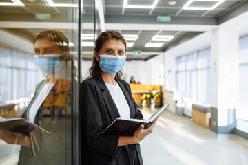 Fototapeta na wymiar Young business woman with a notebook in her hands stands in the office corridor wearing medical mask to protect from coronavirus during epidemic. Health safely at work concept.