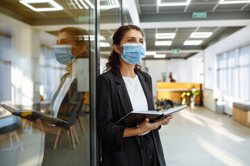 Young business woman with a notebook in her hands stands in the office corridor wearing medical mask to protect from coronavirus during epidemic. Health safely at work concept.