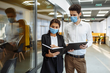 Businesspeople discuss workring issues during pandemic of coronavirus. Man and woman wearing protective medical masks talking business in the office corridor. Health safety at work concept.