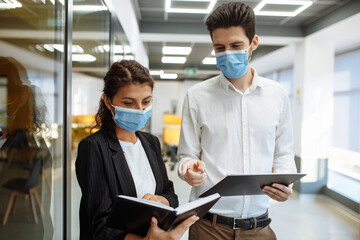 Businesspeople discuss workring issues during pandemic of coronavirus. Man and woman wearing protective medical masks talking business in the office corridor. Health safety at work concept.