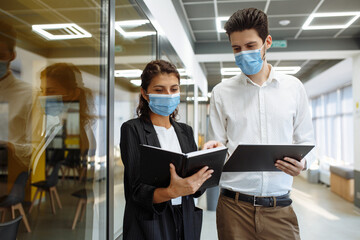 Businesspeople discuss workring issues during pandemic of coronavirus. Man and woman wearing protective medical masks talking business in the office corridor. Health safety at work concept.