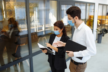 Businesspeople discuss workring issues during pandemic of coronavirus. Man and woman wearing protective medical masks talking business in the office corridor. Health safety at work concept.