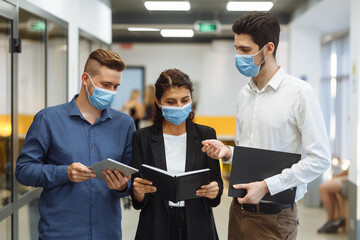 Obraz na płótnie Canvas Three collegues discuss business plan during the break at the office. Coronavirus pandemic measures at work. Group of collegues stay safe wearing medical masks. Health protection concept.