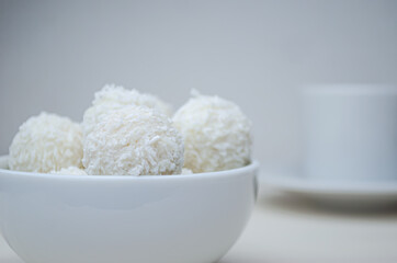 Round candies in a white plate covered with coconut shavings on a white background and with a Cup of espresso coffee.