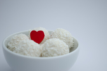 Round candies in a white plate covered with coconut shavings on a white background and with a red heart.