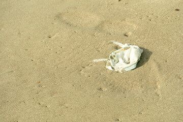 White face medical mask is left abandoned on sand beach. Garbage polluting the natural environment