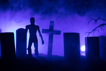 Fototapeta premium Scary view of zombies at cemetery dead tree, moon, church and spooky cloudy sky with fog, Horror Halloween concept with glowing pumpkin. Selective focus