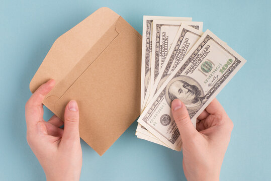 First person above top overhead view photo of female woman hands holding stack pile of american dollars and craft paper envelope isolated over blue color pastel background