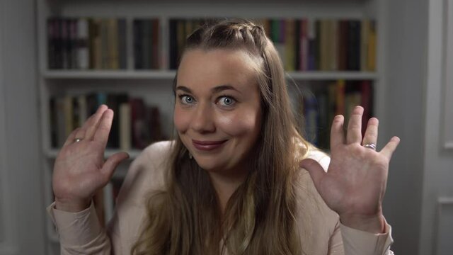 Portrait of caucasian woman who is throwing up her hands and shaking shoulders. Young lady feels perplexity and misunderstanding. She sits in room on bookshelf background. Theme of person's emotions.