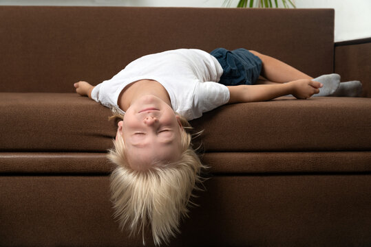 Blond boy lies on couch with his head hanging. Child is tired. Wrong sleeping position of child