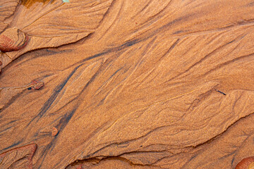 The close-up and abstract texture of patterns of the water on the sand the beach. The red sand washes away, creating grooves for water to flow back and forth across the beach.