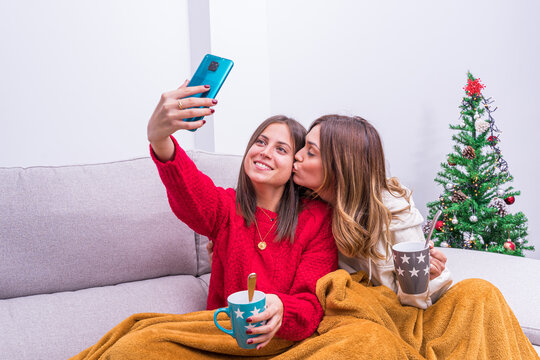 Young lesbian couple drinking coffee, taking pictures and having fun by the Christmas tree. LGBT couple concept, relaxation and home life.