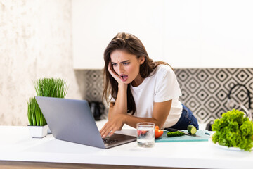 Happy young woman enjoying cooking looking for a recipe on the laptop in the kitchen Laptop on kitchen table and cooking girl. Food blogger concept