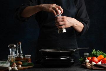 The professional chef in black uniform pours spices in pan with cooking meal on the stove. Backstage of preparing grilled meat for dinner on dark blue background. Food concept. Frozen motion.