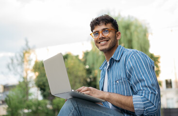 Portrait of handsome Indian copywriter using laptop computer, typing, working freelance project online, sitting outdoors. Smiling asian student studying, learning language, online education concept - obrazy, fototapety, plakaty