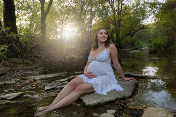 Fototapeta premium Young pregnant lady sitting on rock in forest creek hand on maternity belly beautiful outdoor portrait 
