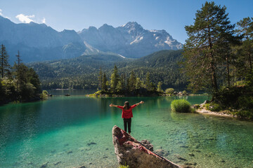 Girl in red jacket standing on a rock and spreading arms, watching a beautiful view over lake...