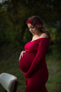 Expecting young lady looking down holding pregnant belly outdoor portrait wearing red gown and flowers in her hair 