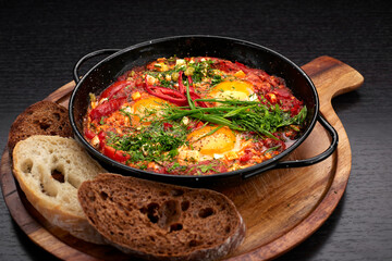 shakshuka on a frying pan and a wooden board, with green onions, bell peppers and croutons, on a dark background