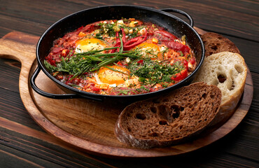 shakshuka on a frying pan and a wooden board, with green onions, bell peppers and croutons, on a wooden background