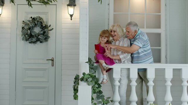 Senior ouple sitting with granddaughter in porch at home. Using mobile phone for online video call