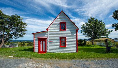 Side view of a vintage cottage under blue sky and clouds. The small white wooden building has two double-hung windows with red trim. There are green grass and a gravel path. 