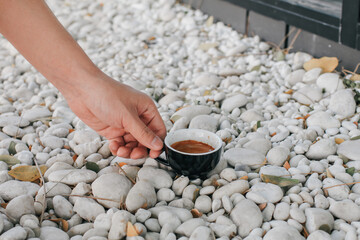 Hand pick up a black cup of hot coffee on white rock in garden floor. Background and texture of white pebbles stone. Take a break from work.
