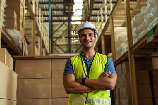 Portrait of young Indian industrial worker with arm folded working in logistic industry indoor inside factory warehouse. Smiling happy man in hard hat looking at camera arms crossed at depot