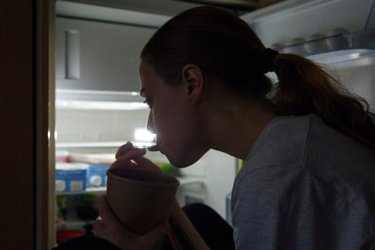 Woman standing near fridge and eating sweet food at night