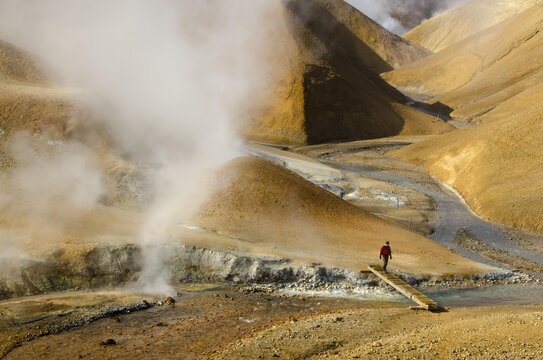 Hiker on a bridge over a steaming creek on a hiking trail, hot springs and the snow-capped Rhyolite Mountains, Hveradallir high temperature area, Kerlingarfjoell, highlands, Iceland, Europe