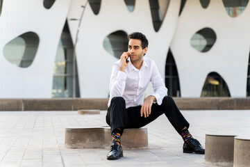 Serious businessman talking over smart phone while sitting on seat against built structure in city