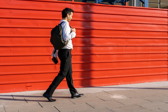 Businessman carrying backpack while walking on footpath by red wall in city