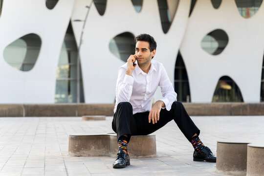 Serious businessman talking over smart phone while sitting on seat against built structure in city