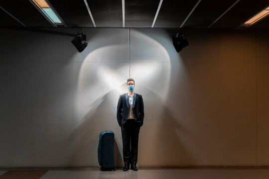 Male professional wearing mask while standing with suitcase against wall at station