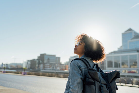Young woman with backpack standing against clear sky in city during sunny day