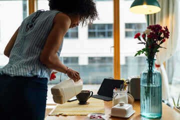 Young woman pouring milk in coffee cup on table at home