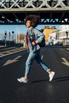 Cheerful young woman with afro hair running on street in city during sunny day