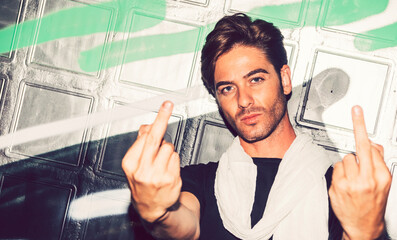 Close-up of handsome man showing middle fingers against graffiti wall at night