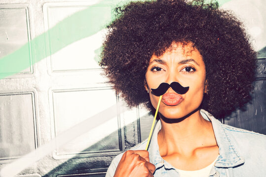 Close-up of woman puckering lips while holding fake mustache on face against wall at night