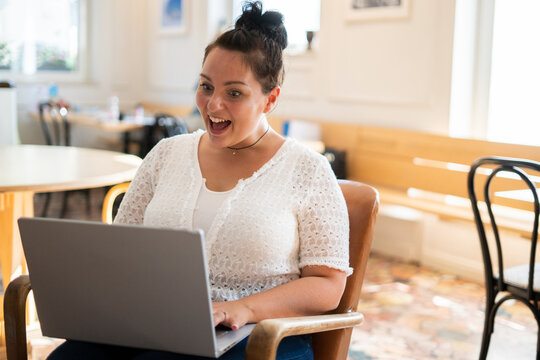 Shocked young woman with mouth open using laptop while sitting on chair in cafe