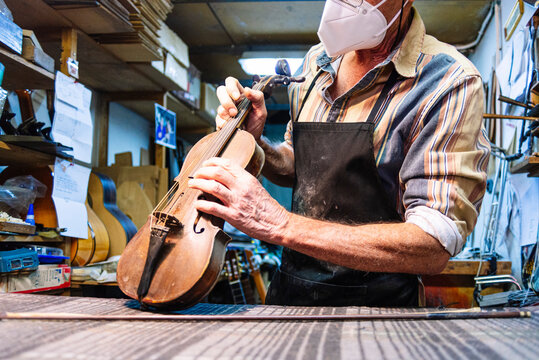 Craftsman with face mask examining string of violin on workbench at workshop