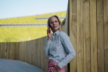 Beautiful blond female professional looking away while talking on smart phone against wooden surrounding wall