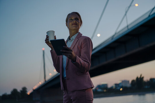 Businesswoman looking away while holding digital tablet and disposable cup against bridge at dusk