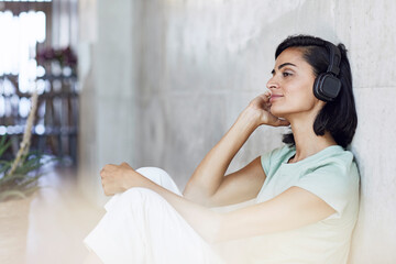 Thoughtful female entrepreneur listening music over headphones while sitting against wall in office