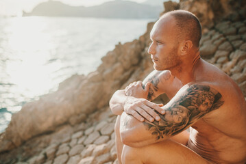 Tattooed nudist sitting on volcanic rocks by the sea
