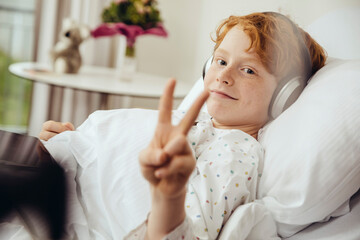 Sick boy lying in hospital making victory sign, wearing head phones