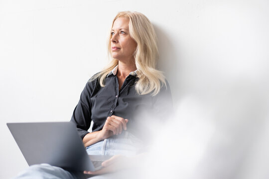 Thoughtful Businesswoman With Laptop Sitting Against White Wall In Office