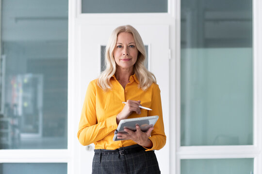 Confident female entrepreneur holding digital tablet while standing against wall in home office