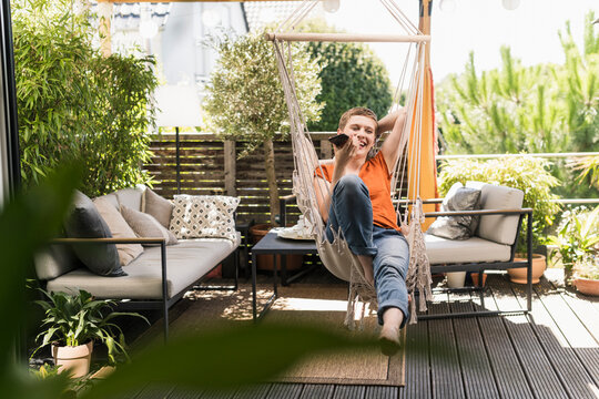 Cheerful mid adult woman talking over smart phone while swinging in porch seen through indoors